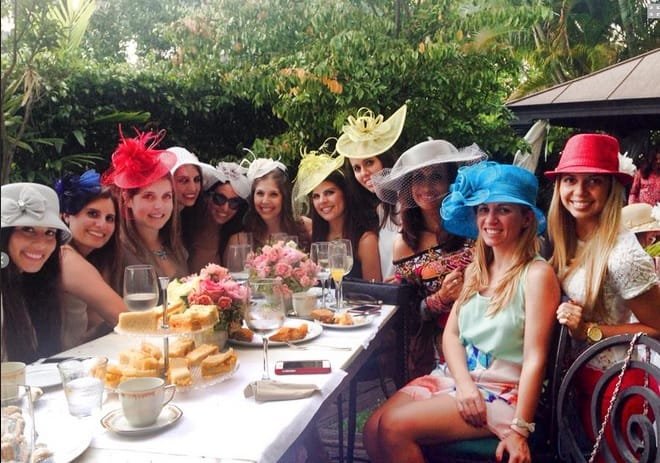 A group of women in hats sitting at a table with food and drinks outdoors.